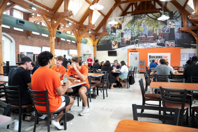 Students sit and talk in a busy university dining hall with wooden beams and a large mural on the back wall.