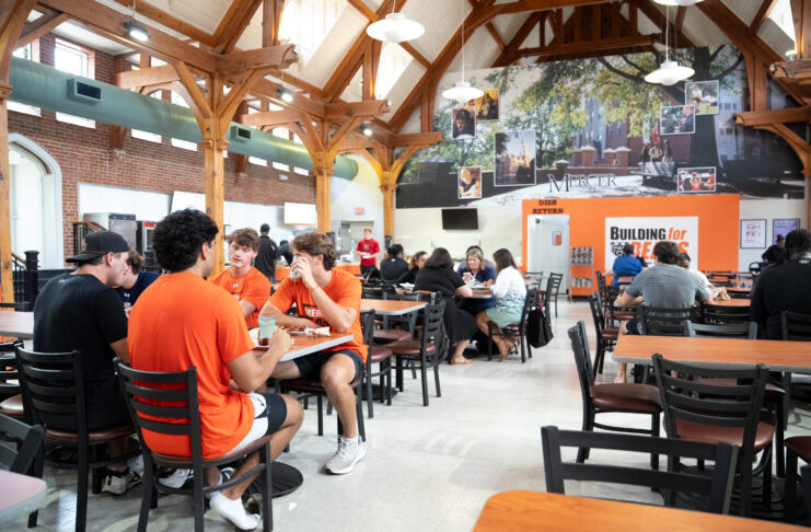Students sit and talk in a busy university dining hall with wooden beams and a large mural on the back wall.