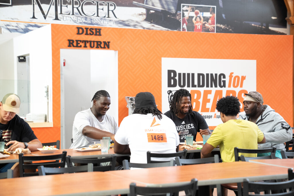 Six men sit and eat together at a cafeteria table, with signs and food trays visible in the background.