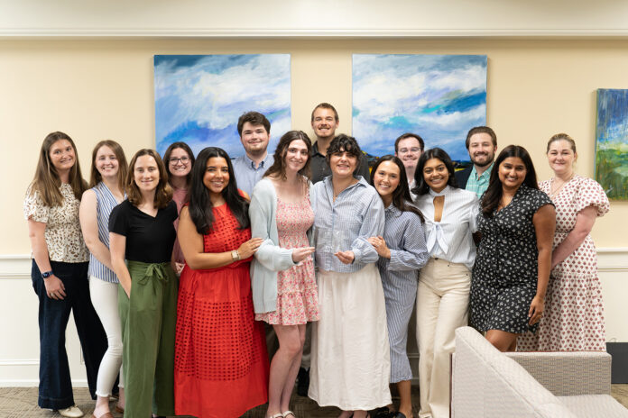A group of fourteen people stand indoors, smiling at the camera, in front of two abstract blue and green paintings.