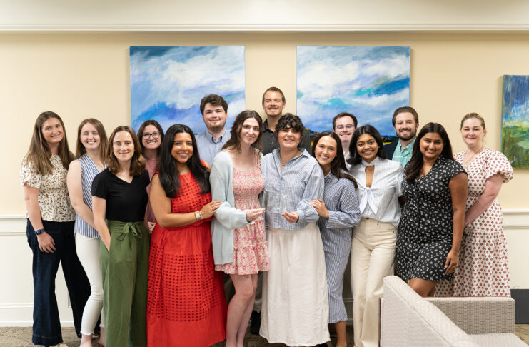 A group of fourteen people stand indoors, smiling at the camera, in front of two abstract blue and green paintings.