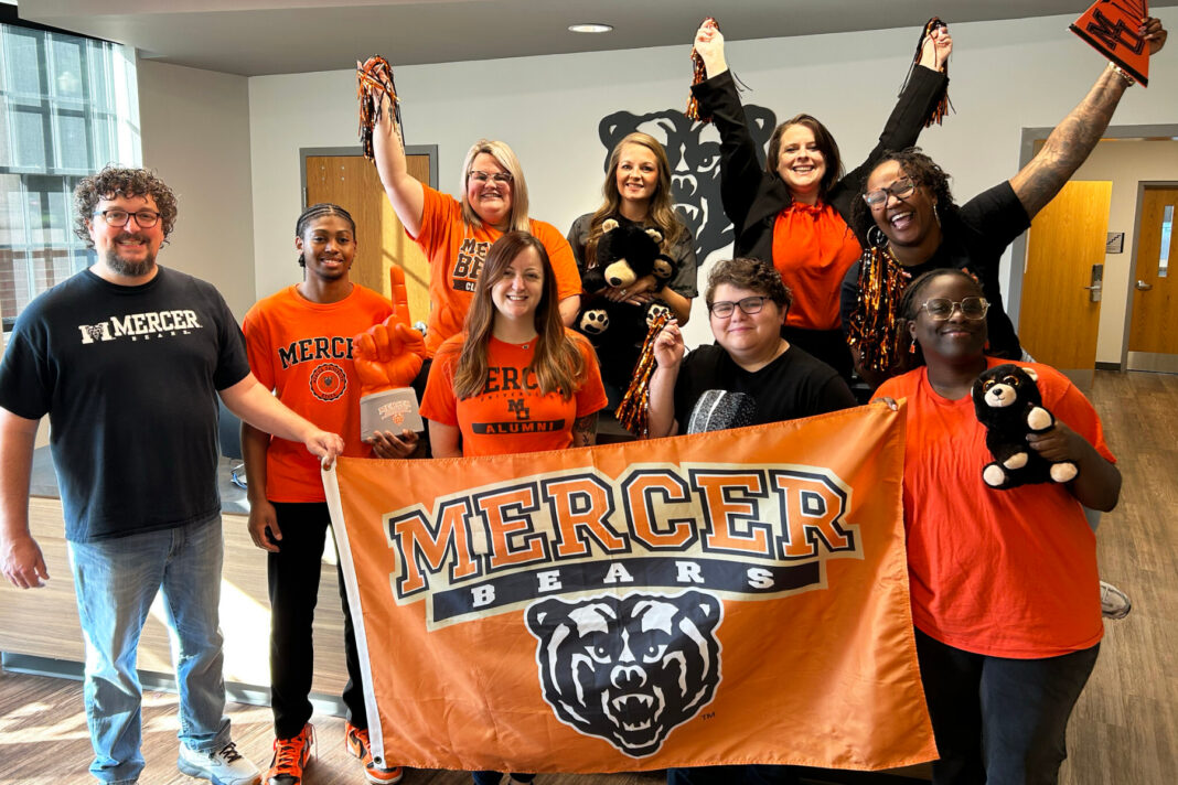 Group of people in Mercer Bears gear holding a school banner, pom-poms, and teddy bears, posing indoors and smiling.