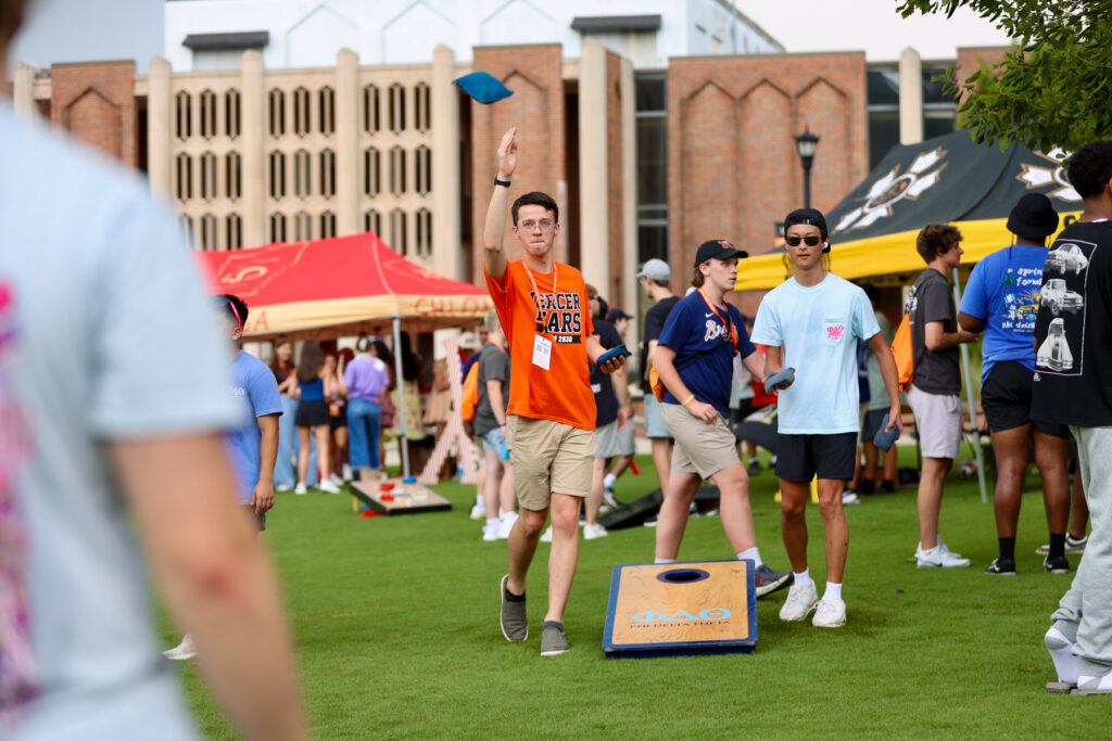 A young man tosses a bean bag while playing cornhole at an outdoor event with other people and tents in the background.