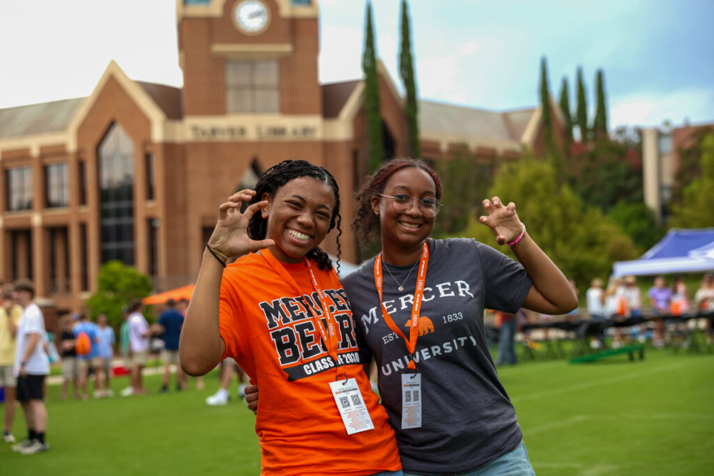 Two students wearing Mercer University shirts pose and smile on a grassy campus lawn with a clock tower in the background.