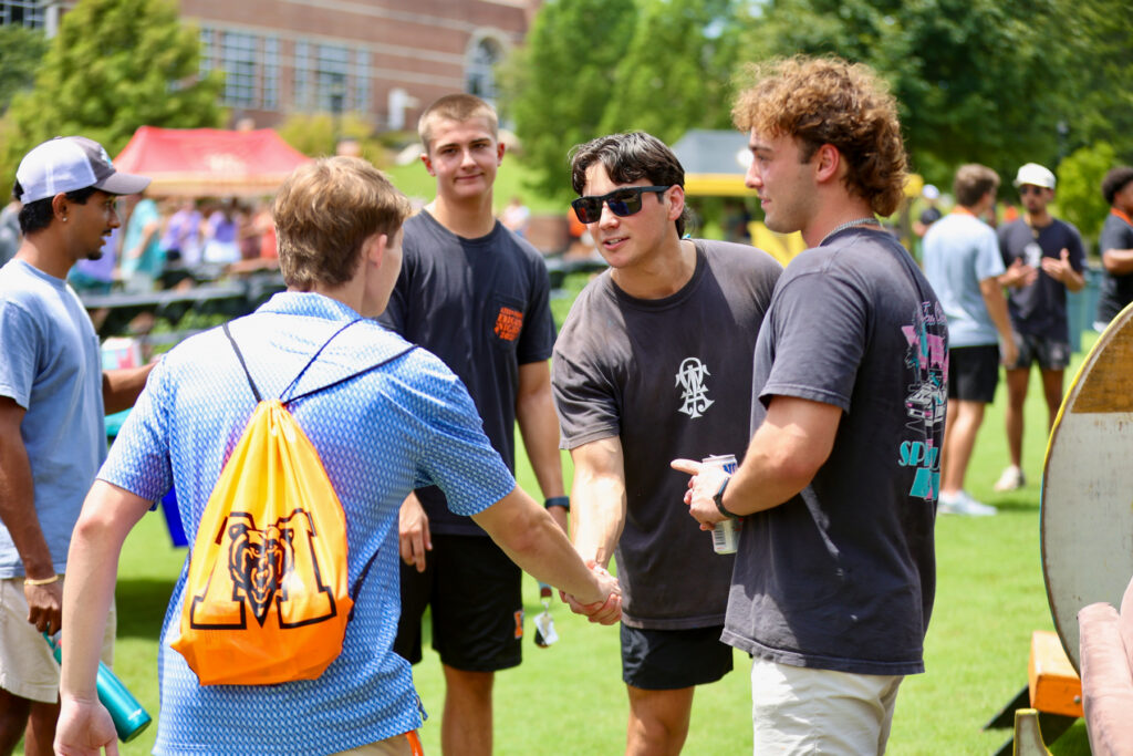 Four young men greet each other outdoors on a sunny day; one carries an orange drawstring backpack.