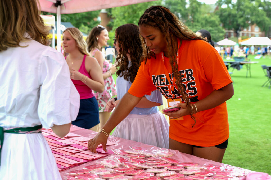A woman in an orange shirt selects a cookie from a table at an outdoor event with other people in the background.