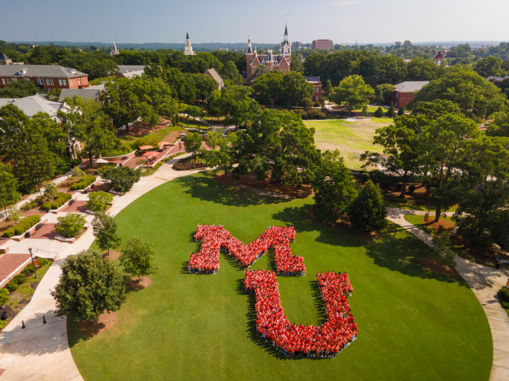 Students form a large MU on a campus lawn, with historic university buildings and trees in the background on a sunny day.