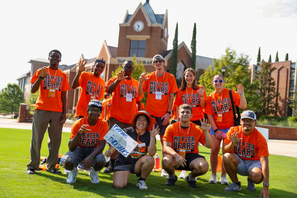 A group of students in orange Mercer Bears shirts pose and smile on a campus lawn in front of a brick building.
