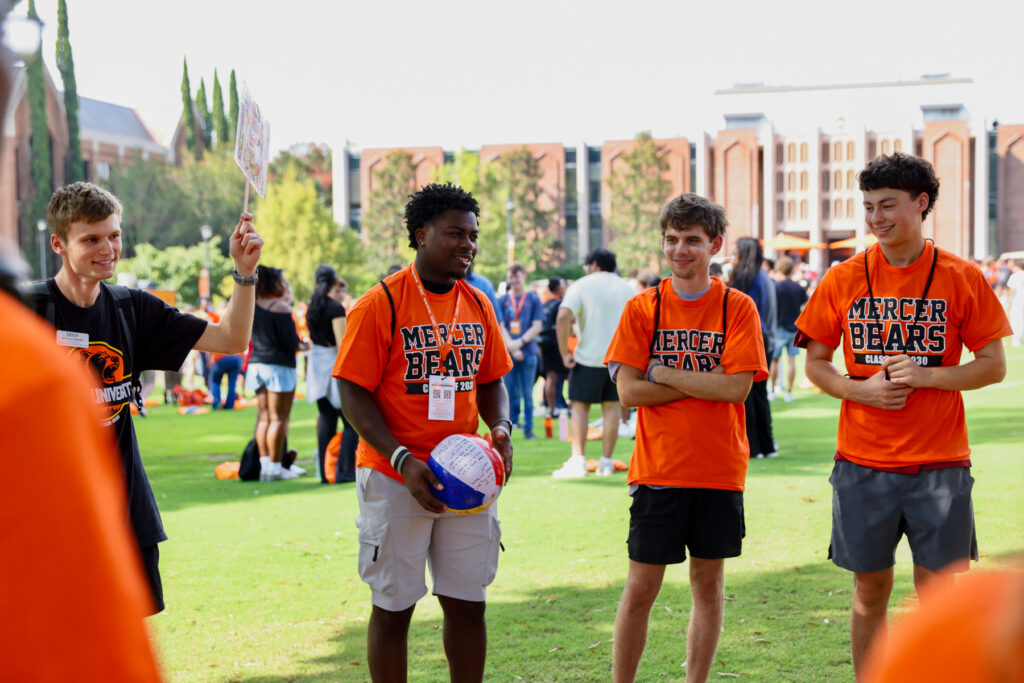 Four students wearing Mercer Bears shirts stand together on a lawn during an outdoor campus event.