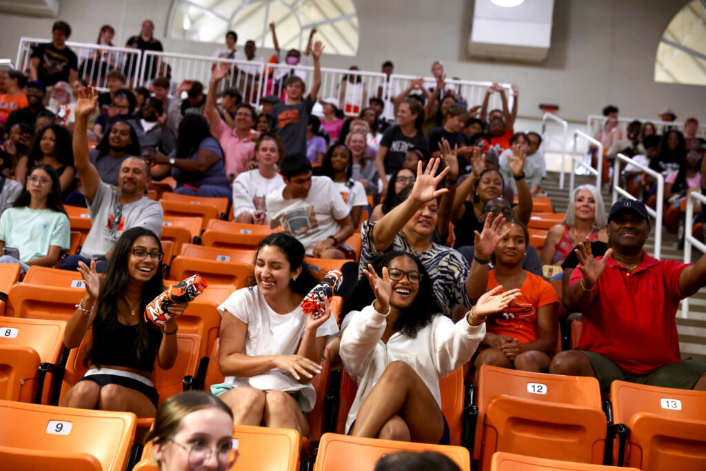 A diverse group of people sit in orange stadium seats, smiling and waving toward the camera.