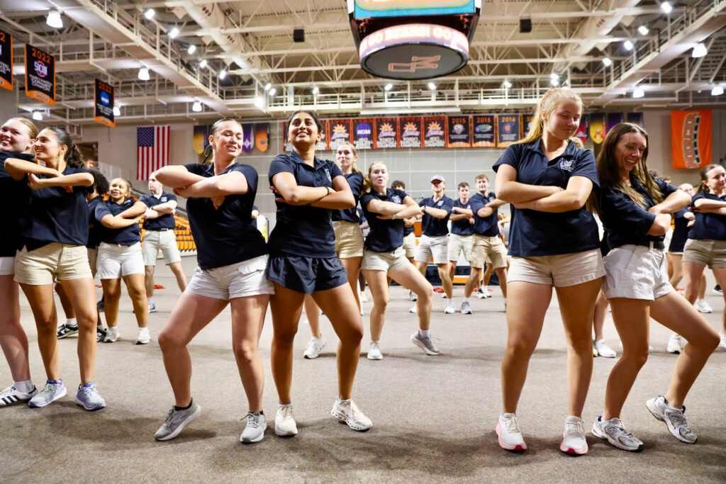 A group of students in matching shirts and shorts perform a synchronized dance routine in a gymnasium.