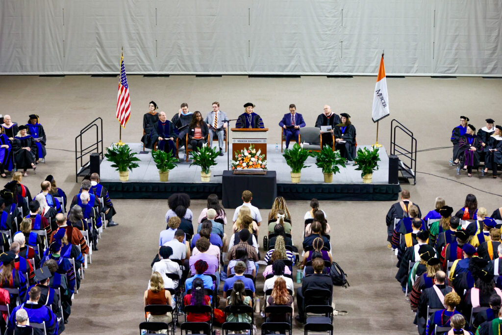 Convcation ceremony with faculty on stage, audience seated facing forward, and flags displayed on each side of the stage.