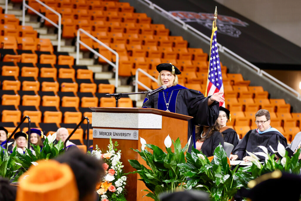 Wearing academic regalia, Mercer President Penny L. Elkins, Ph.D., speaks at a podium during convocation in Hawkins Arena.