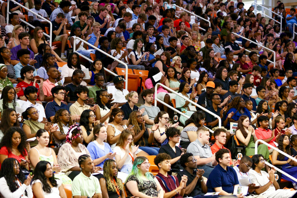 Large audience seated in Hawkins Arena bleachers, watching convocation and applauding, with many people visible.