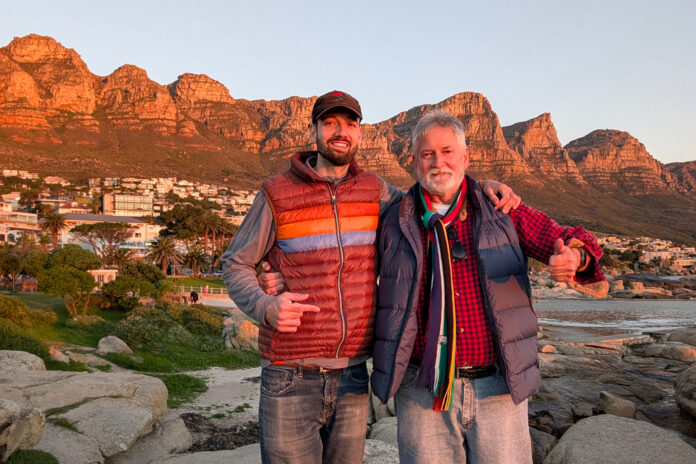 father son 2 crop Two men in jackets stand on rocks, smiling, with mountains and houses in the background at sunset.