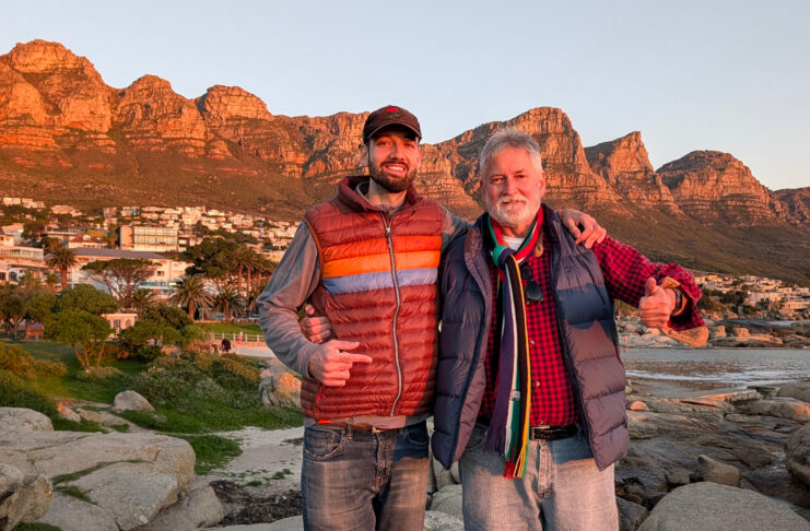 Two men in jackets stand on rocks, smiling, with mountains and houses in the background at sunset.