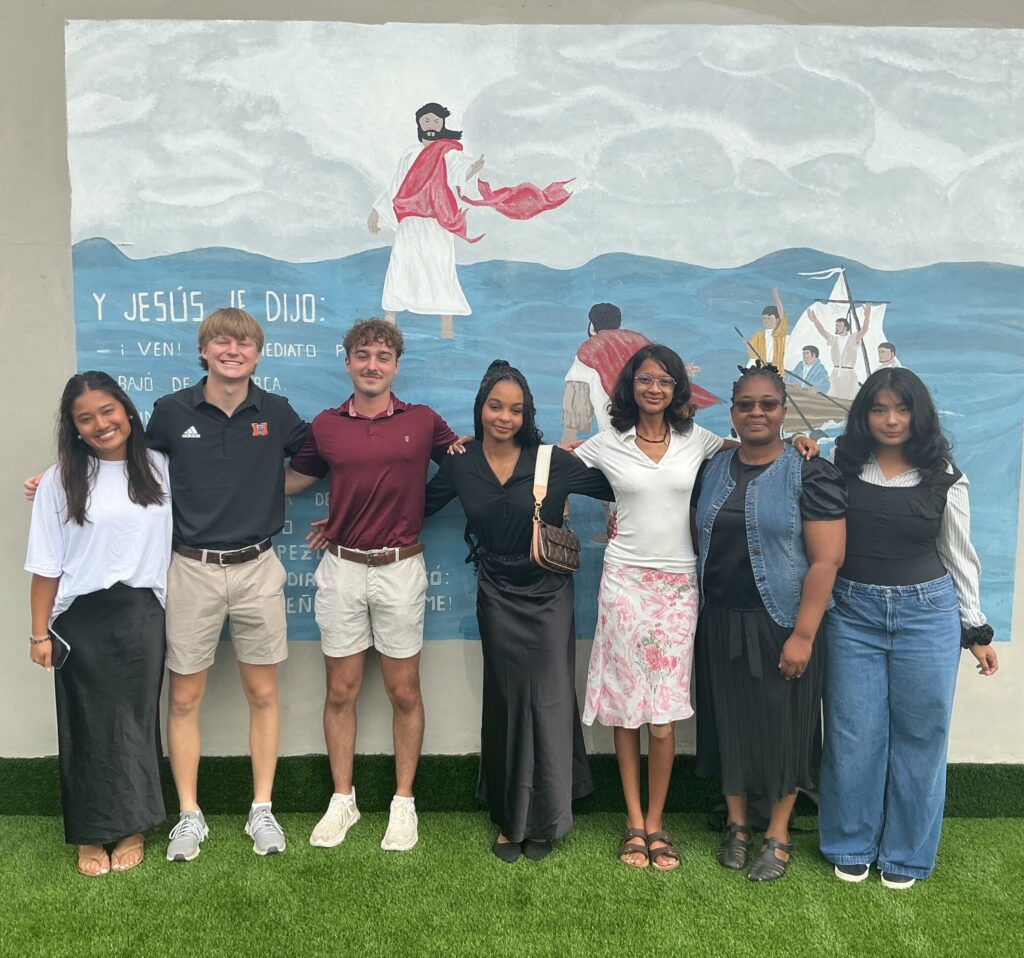 Seven people stand in a row and smile in front of a painted mural on a wall with grass beneath their feet.