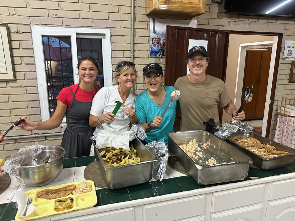 Four people stand behind a counter serving food from metal trays in a kitchen or cafeteria setting.