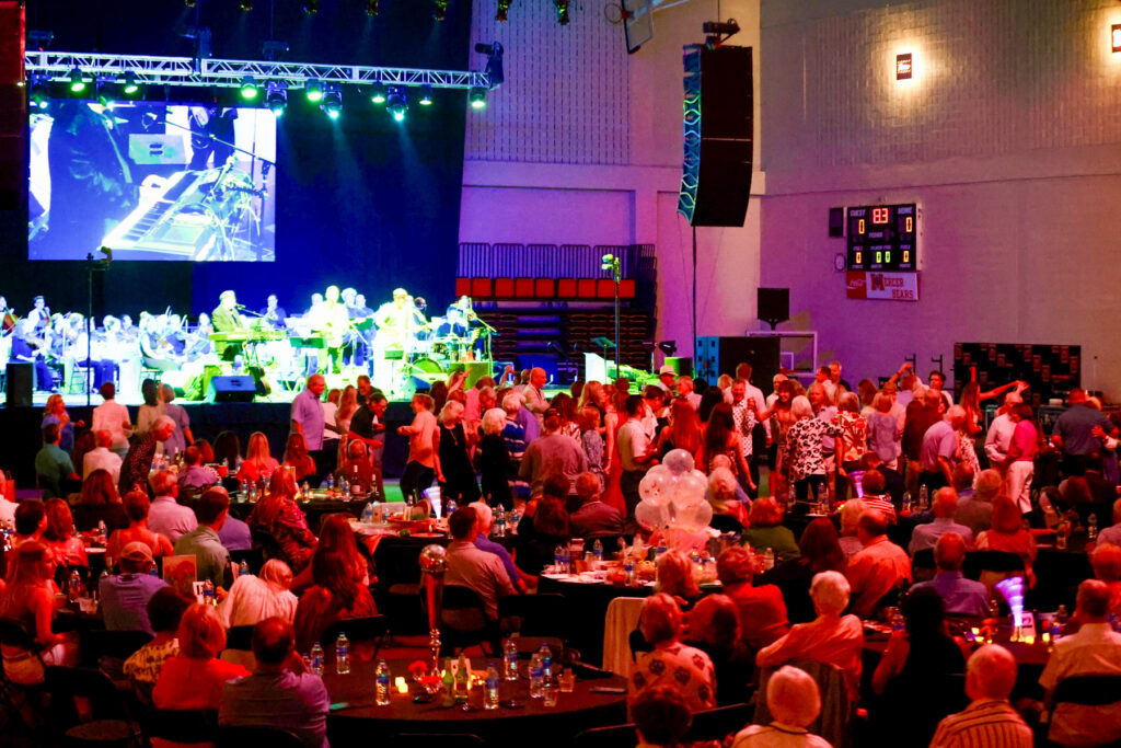 Large crowd seated and standing at tables in a gymnasium, watching a live band perform on a stage with bright lights.