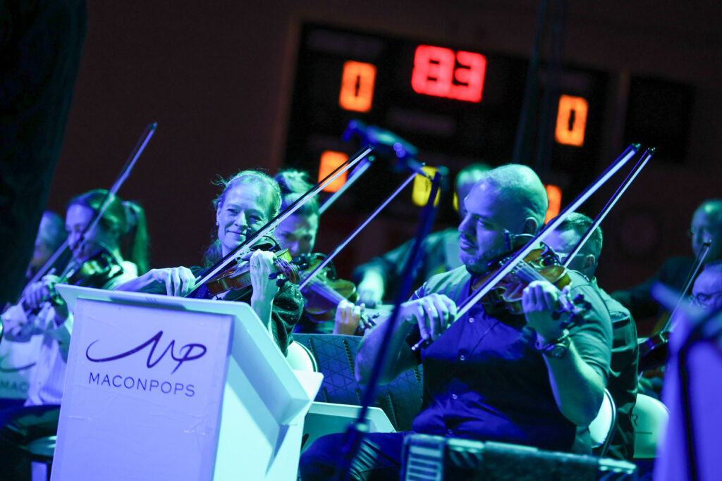 Musicians play violins at a Macon Pops event, with a digital scoreboard visible in the background.