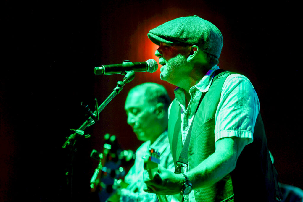 Two musicians perform on stage, one singing into a microphone and playing guitar, both under green stage lighting.