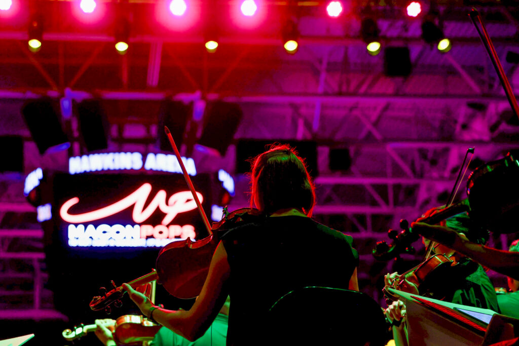 Musicians play string instruments on stage under pink lights, with a Macon Pops sign visible in the background.