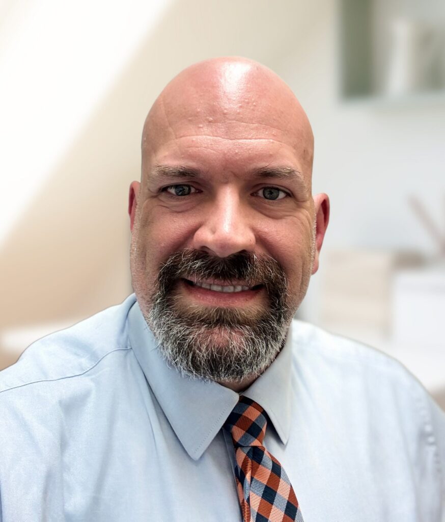 Bald man with a beard and mustache, wearing a light blue shirt and checkered tie, smiling indoors.