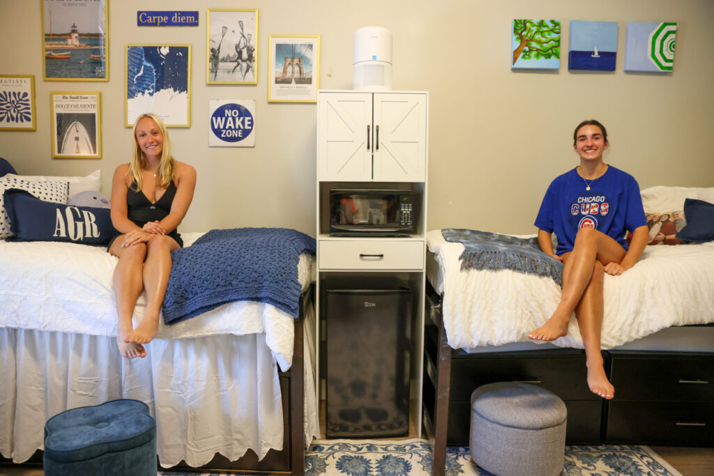 Two people sit on separate beds in a neatly decorated dorm room, with a microwave and fridge between them.