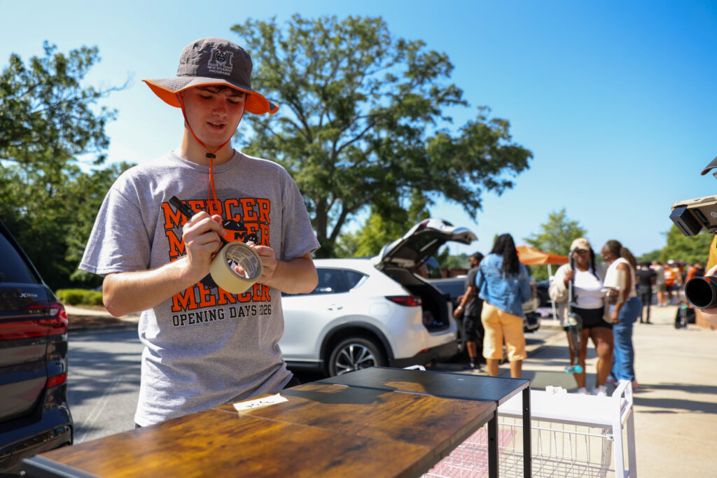 A person in a Mercer University shirt labels an item outdoors while people unload cars in the background.