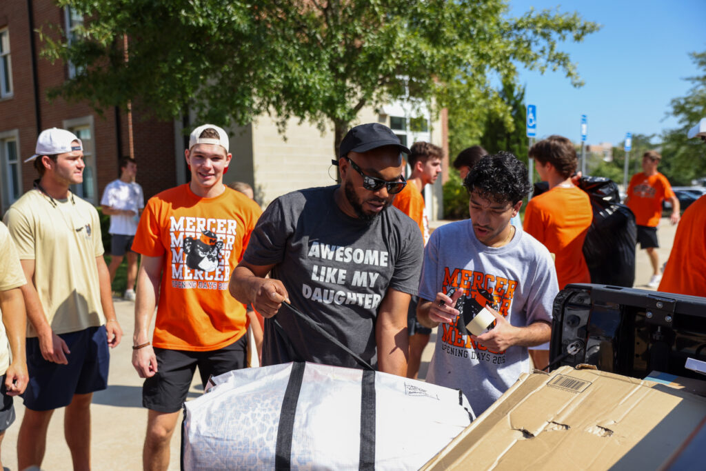 Group of people unloading boxes and bags from a car outside a building on a sunny day.