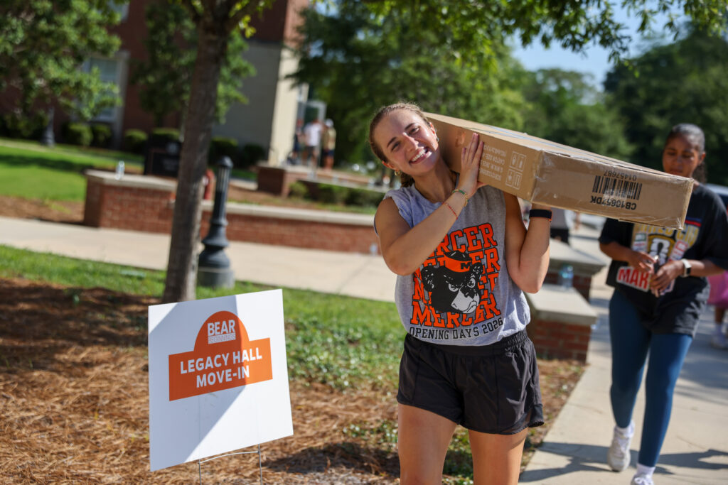 Smiling person carrying a box near a Bear Legacy Hall Move-In sign on a sunny day.