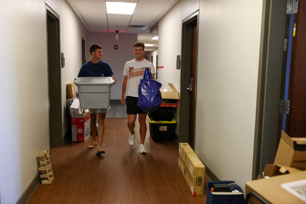 Two people carry bins and bags down a hallway lined with boxes and items, possibly moving into a dorm or apartment.