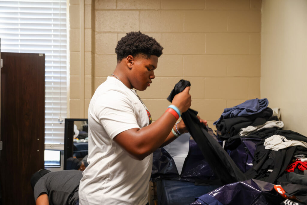 A person folds clothes in a room with beige walls, a bed, and clothes piled on the bed and nearby furniture.