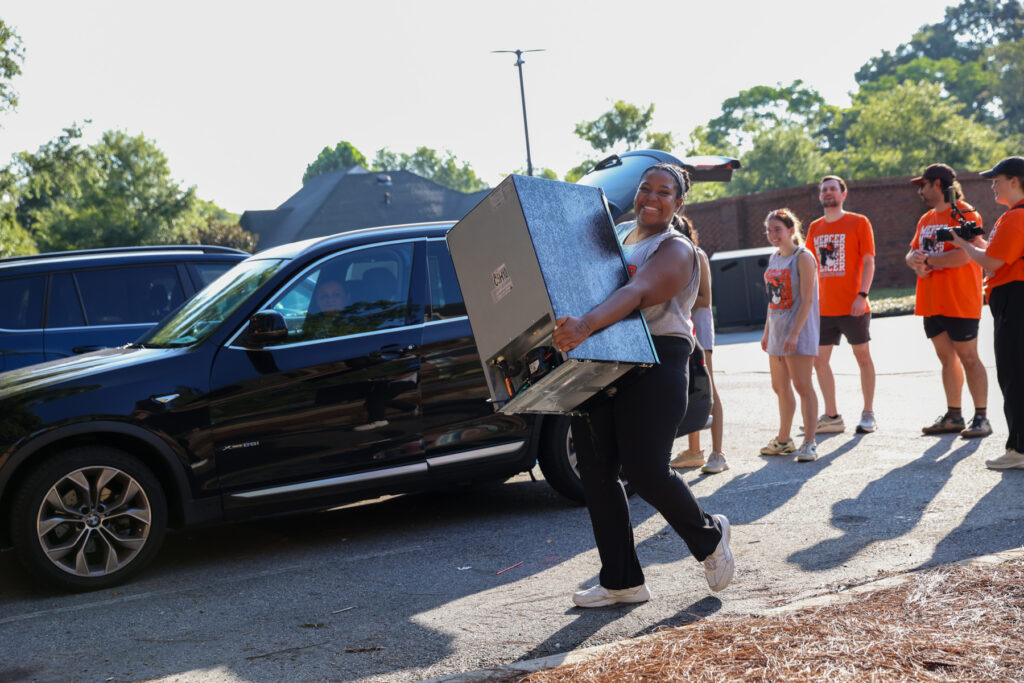 Person carrying a mini fridge from a car, with a group of people standing and watching outside.
