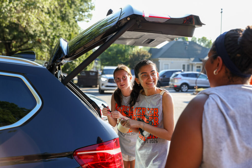 Two teenage girls unload items from an SUV trunk while talking to a woman in a parking lot on a sunny day.