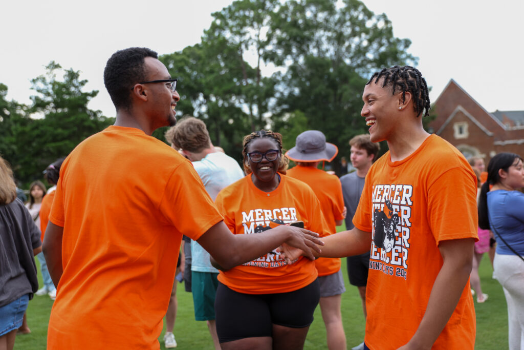 Two people in orange shirts shake hands outdoors, while another person in similar attire smiles in the background.