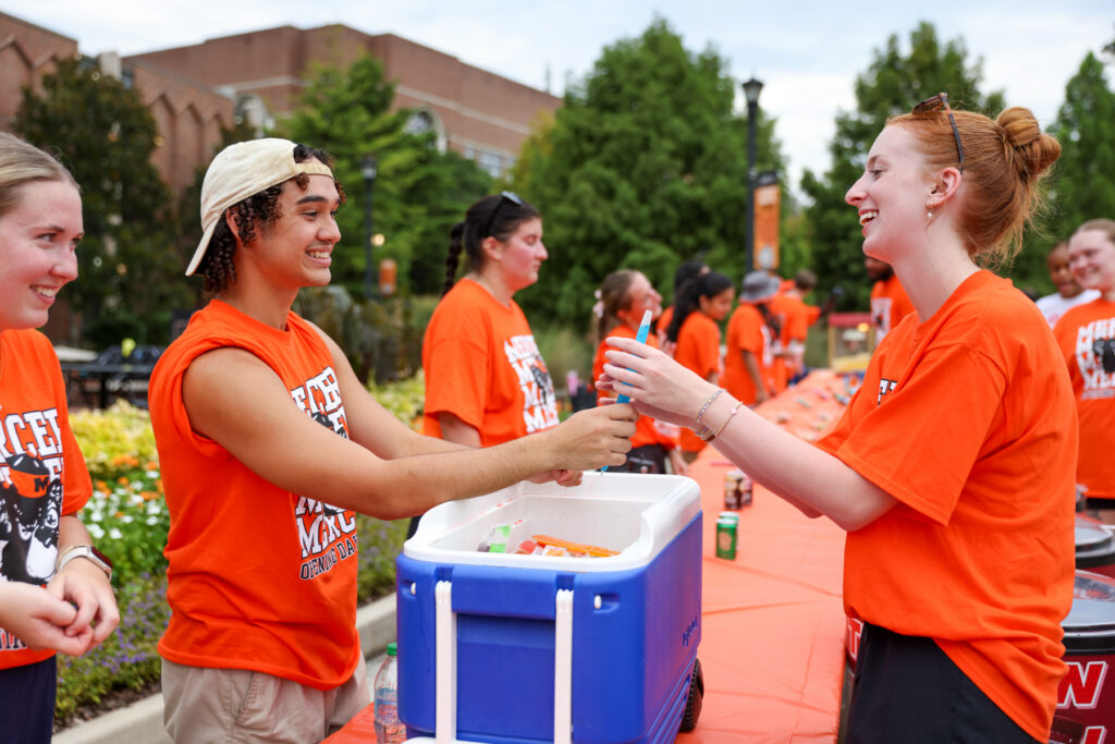 A group of people in orange shirts stand by a table while one person hands a can to another at an outdoor event.