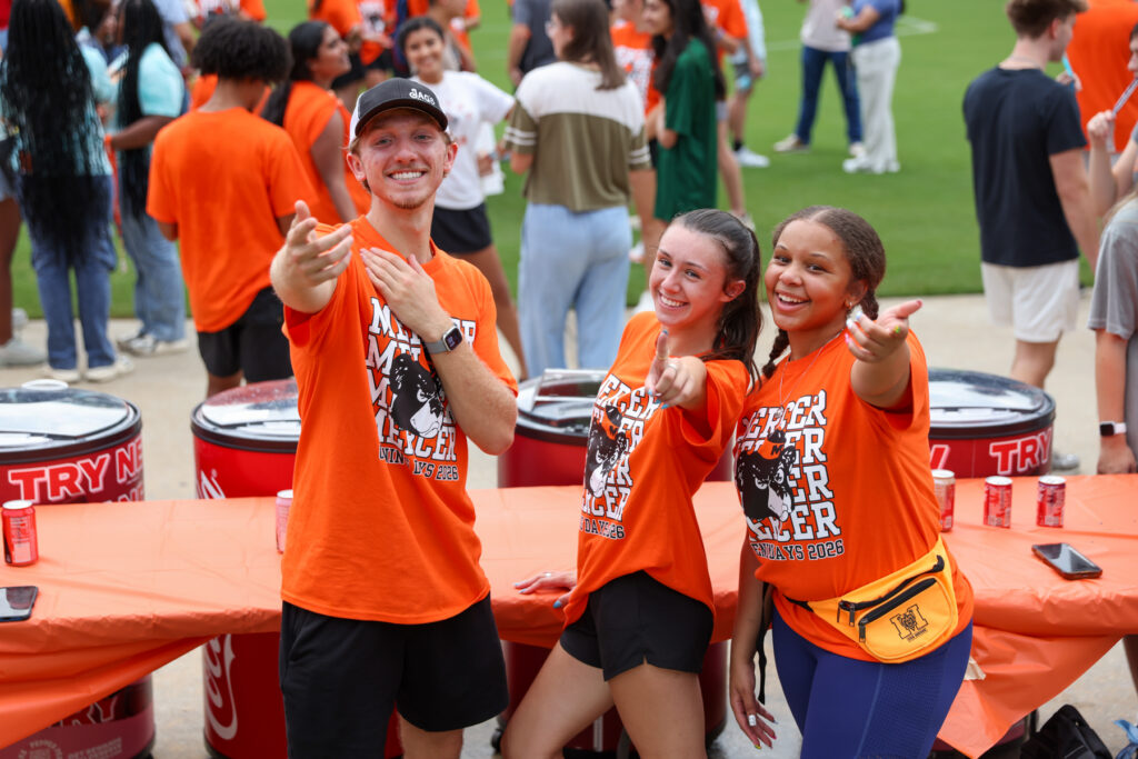 Three smiling people in orange shirts pose and point at the camera at an outdoor event with others in the background.