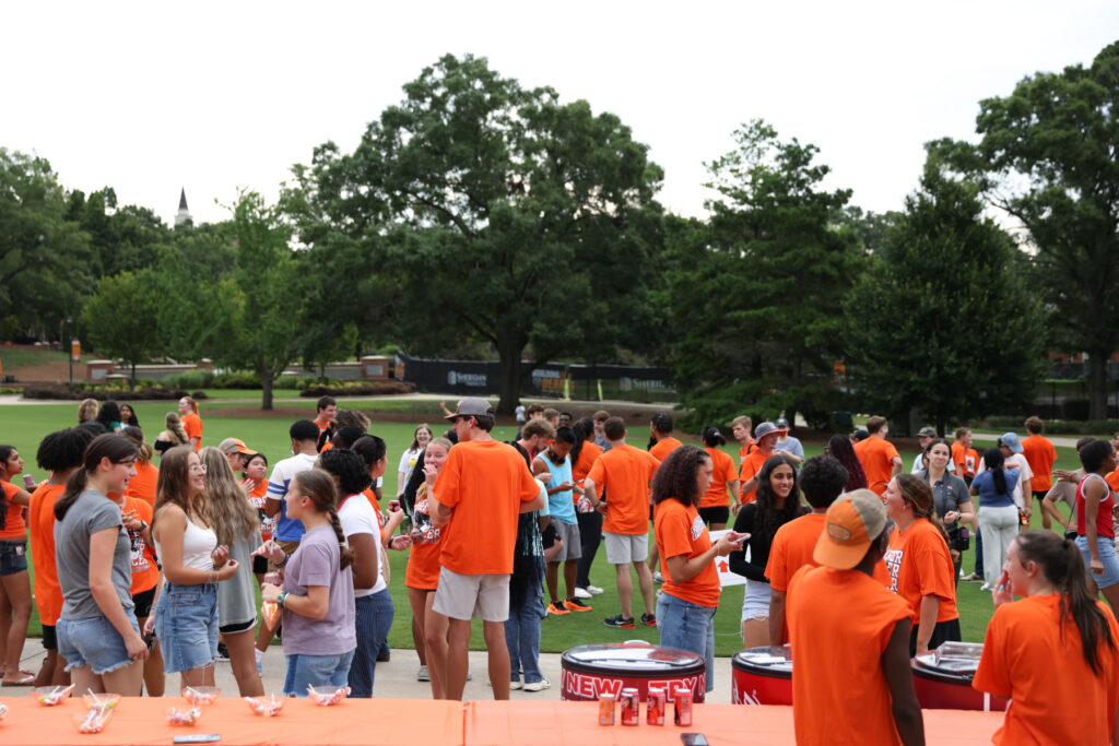A group of people, many in orange shirts, gather outdoors near tables with drinks and snacks on a sunny day.