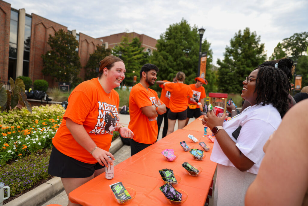 People interact at an outdoor event table with snacks and drinks, wearing orange shirts, on a campus walkway.