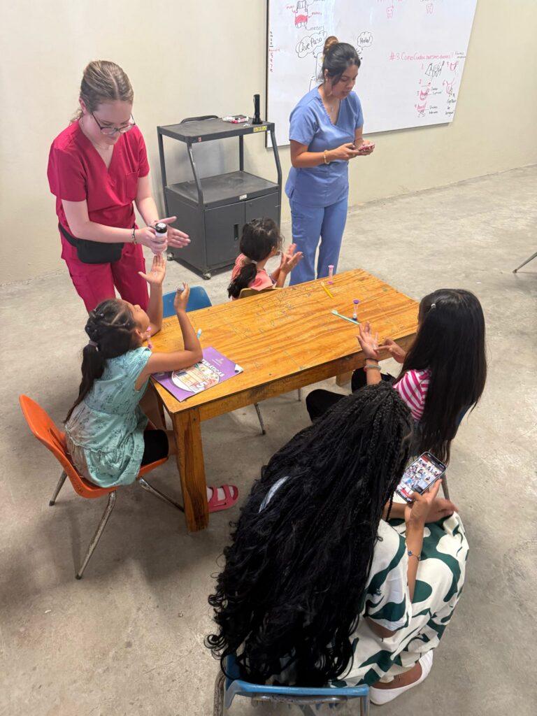 Two women supervise four children sitting at a table with crafts; one woman pours sanitizer on a childs hands.