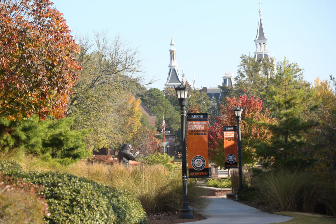 Tree-lined campus walkway with lampposts, banners, and distant view of historic building with towers on a sunny day.