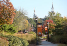 Mercer rises in nation’s top university rankings by U.S. News & World Report Tree-lined campus walkway with lampposts, banners, and distant view of historic building with towers on a sunny day.