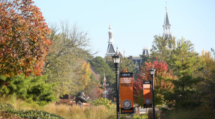 Mercer rises in nation’s top university rankings by U.S. News & World Report Tree-lined campus walkway with lampposts, banners, and distant view of historic building with towers on a sunny day.