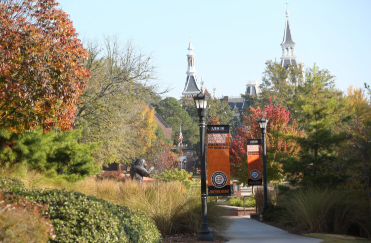 Tree-lined campus walkway with lampposts, banners, and distant view of historic building with towers on a sunny day.