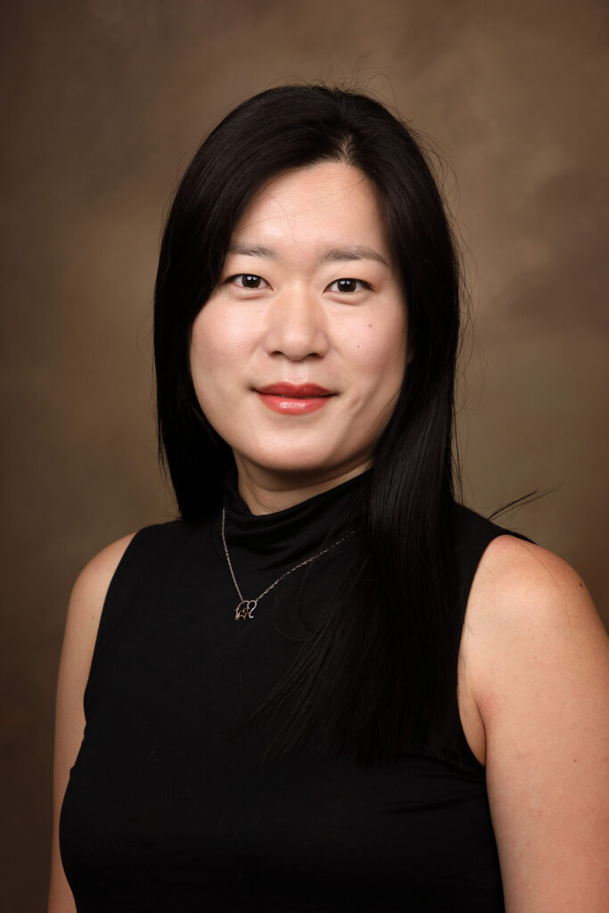 Woman with long black hair wearing a black sleeveless top poses against a plain brown background, facing the camera.