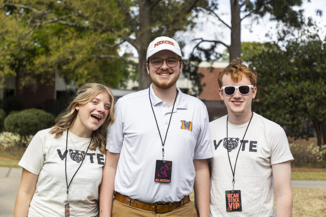 Three smiling young adults stand outdoors wearing event lanyards and Mercer-themed shirts, with trees in the background.