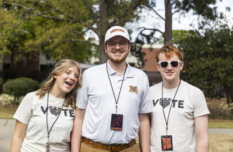 Three smiling young adults stand outdoors wearing event lanyards and Mercer-themed shirts, with trees in the background.