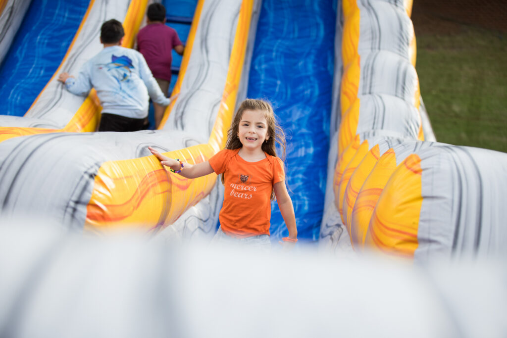 A young girl in a Mercer shirt smiles while climbing an inflatable slide as other children play behind her.
