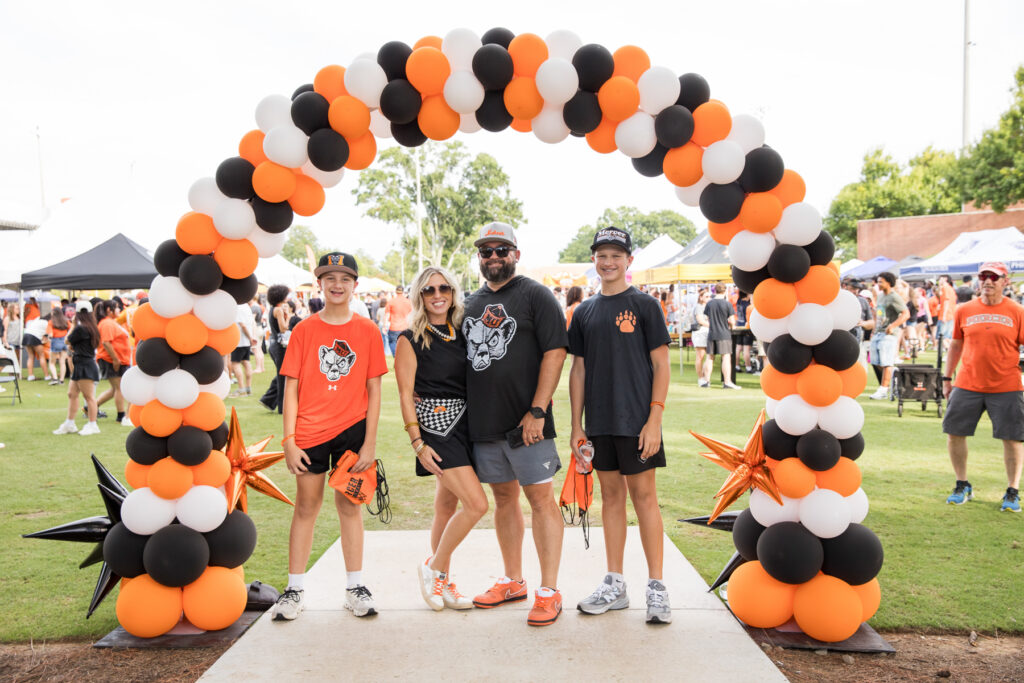 Four people pose under a black, white, and orange balloon arch.